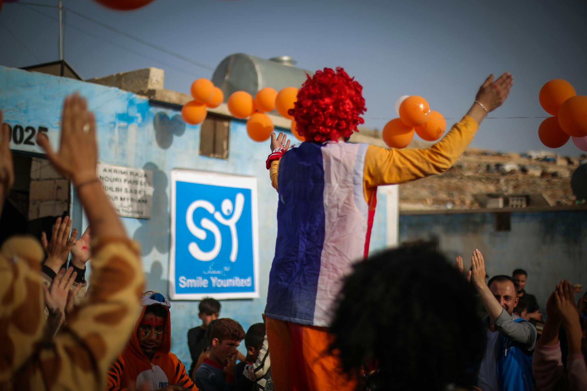 Colorful street performance featuring a clown entertaining a lively crowd in Idlib, Syria.